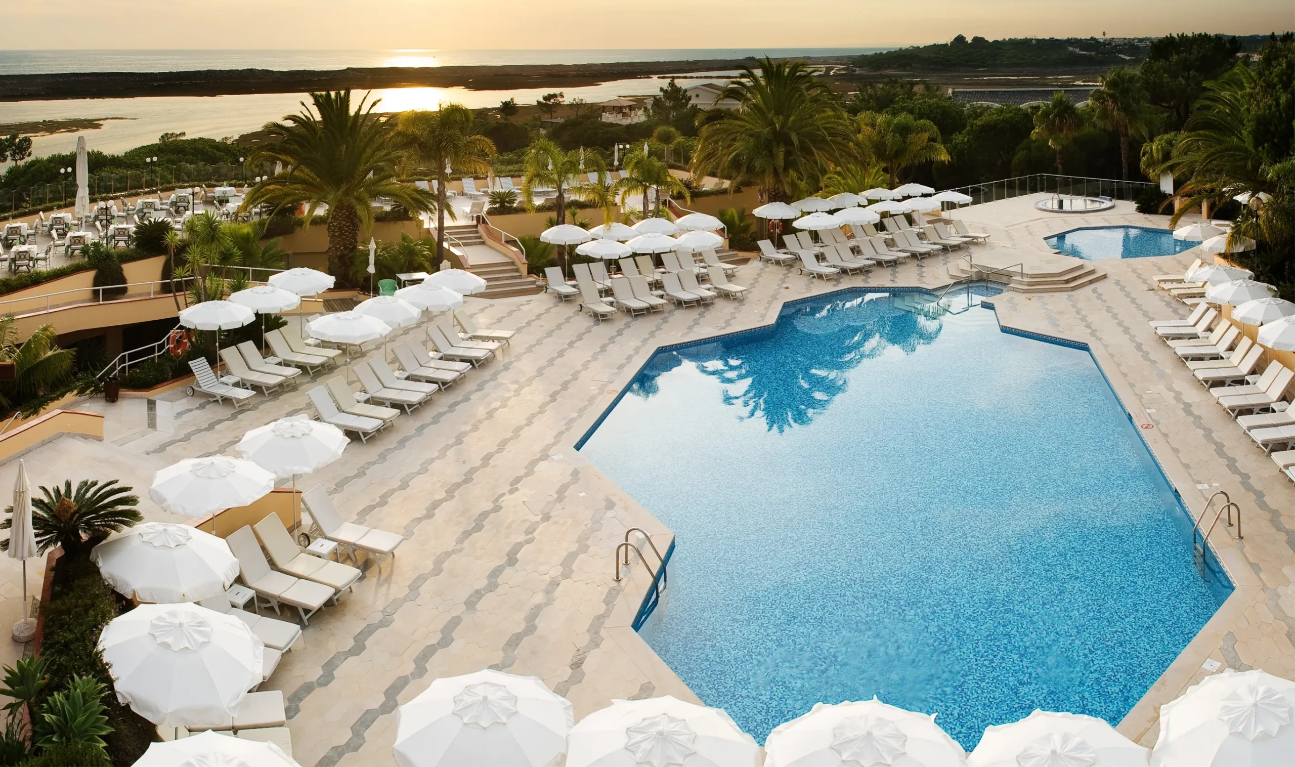 Der große Außenpool des Quinta do Lago Hotels ist von weißen Liegestühlen und Sonnenschirmen umgeben, im Hintergrund erheben sich Palmen und bieten einen malerischen Blick auf das Wasser und den Sonnenuntergang.