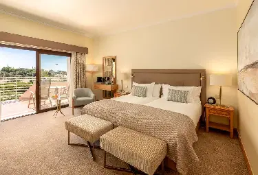 Helles Hotelzimmer im Quinta do Lago Hotel mit zwei Einzelbetten, einer Einrichtung in sanften Beigetönen, einem Schreibtisch, einem Sessel, Kunstwerken an den Wänden und einem Balkon mit Sitzgelegenheiten im Freien und Blick auf eine grüne Landschaft.