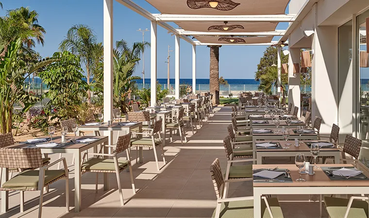 Eine Restaurantterrasse im Freien mit ordentlich aufgestellten Tischen und Stühlen, im Schatten einer weißen Pergola, umgeben von Pflanzen und mit Blick auf einen Sandstrand und das blaue Meer unter einem klaren Himmel – eine idyllische Kulisse, die die entspannte Eleganz des Hipotels Flamenco perfekt widerspiegelt.