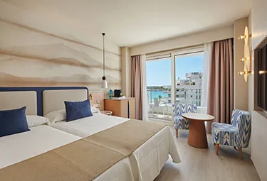Helles Hotelzimmer mit zwei Einzelbetten, in Blau- und Beigetönen gehalten, einem kleinen Tisch mit zwei Stühlen, einem an der Wand montierten Fernseher und einem Fenster, das auf einen Balkon mit Blick auf das Meer und die Gebäude führt – ideal zum Entspannen im Hipotels Flamenco.