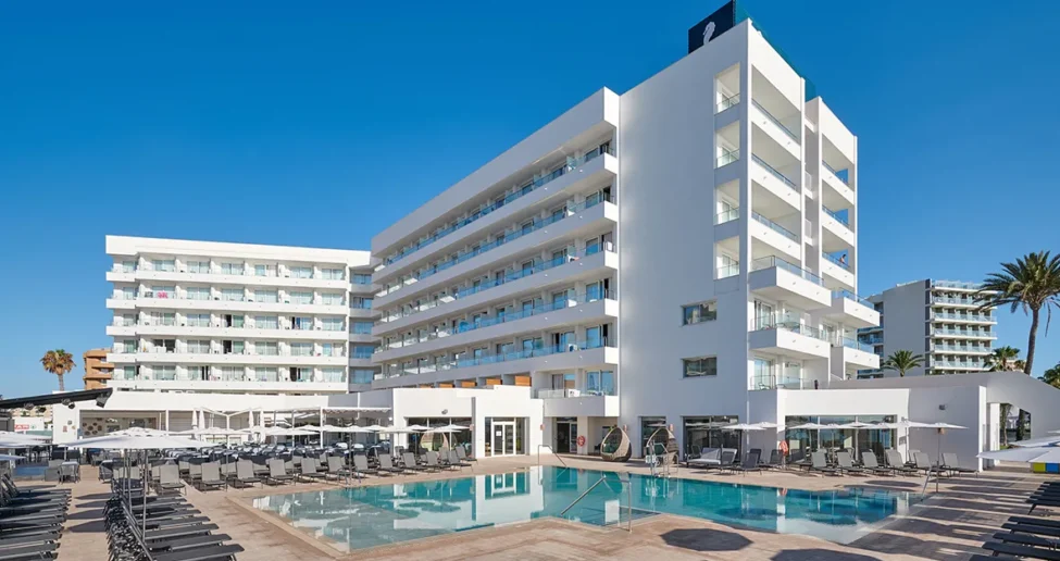 Das moderne, weiße Hotel „Hipotels Flamenco“ mit seinen zahlreichen Balkonen liegt direkt an einem klarblauen Swimmingpool, der von leeren Liegestühlen unter einem strahlend blauen Himmel umgeben ist.