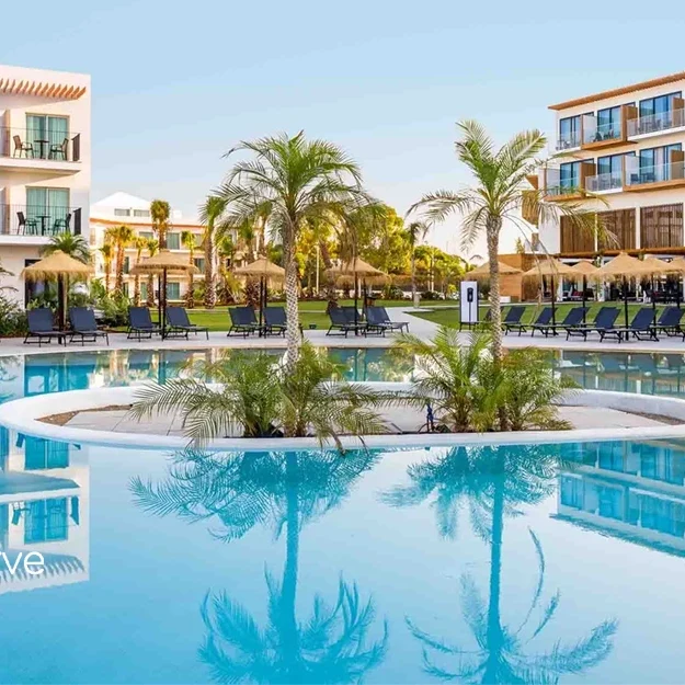 AP-Cabanas-tavira-pool-mitLogo Das moderne Resort AP Cabanas Beach verfügt über einen großen Außenpool, der von Palmen, Liegestühlen und Sonnenschirmen umgeben ist. Zwei mehrstöckige Gebäude mit Balkonen überblicken den Pool. Das Logo von Visit Algarve Portugal befindet sich in der Ecke.