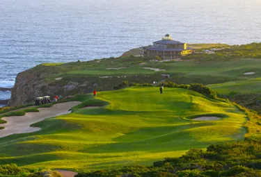 Golfer spielen auf einem üppigen, grünen Golfplatz auf den Klippen über dem Meer, mit einem Clubhaus im Hintergrund und Sonnenlicht, das sich auf dem Wasser spiegelt - ein Erlebnis, das an eine traumhafte Golfreise in Südafrika erinnert.
