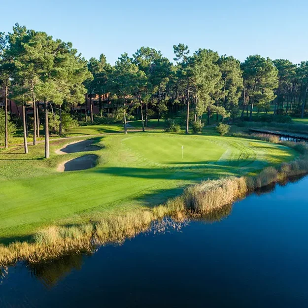 Ein von Bäumen und einem großen Wasserhindernis gesäumtes Golfplatzgrün unter strahlend blauem Himmel, mit Sandbunkern in der Nähe des Grüns und Spiegelungen von Bäumen im Wasser in MARINA CAMBRILS BY URH.
