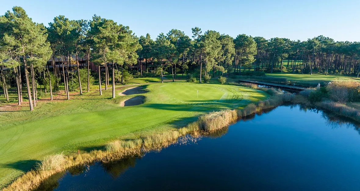 Ein von Bäumen und einem großen Wasserhindernis gesäumtes Golfplatzgrün unter strahlend blauem Himmel, mit Sandbunkern in der Nähe des Grüns und Spiegelungen von Bäumen im Wasser in MARINA CAMBRILS BY URH.