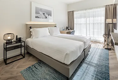 Modernes Hotelzimmer im MARINA CAMBRILS BY URH mit zwei Einzelbetten, weißer Bettwäsche, einem Nachttisch mit Lampe, einem Schreibtisch und einem Stuhl, einem bodentiefen Fenster mit durchsichtigen Vorhängen und einem blau gemusterten Teppich auf Holzfußboden.