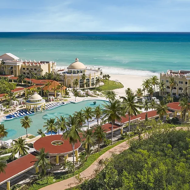 Luftaufnahme des Joia Paraiso by Iberostar Beachfront Resort mit Palmen, einem großen Swimmingpool, Gebäuden mit roten Dächern und weißem Sandstrand mit Blick auf den türkisfarbenen Ozean unter einem klaren blauen Himmel.