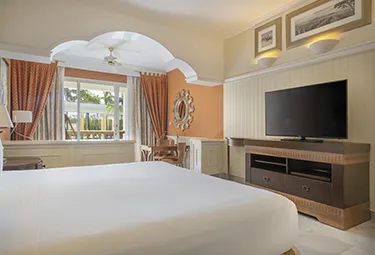 Ein helles Hotelzimmer im Joia Paraiso by Iberostar verfügt über ein großes Bett, einen Flachbildfernseher auf einem Holzständer, einen Schreibtisch mit Stuhl in der Nähe eines Fensters, eine orangefarbene Akzentwand und weiße und orangefarbene Vorhänge. Ein gerahmtes Kunstwerk hängt über dem Fernseher.