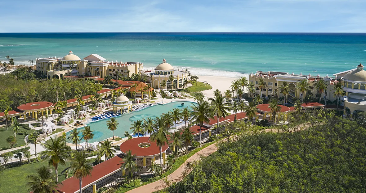 JOIA Paraiso by Iberostar Luftaufnahme des Joia Paraiso by Iberostar Beachfront Resort mit Palmen, einem großen Swimmingpool, Gebäuden mit roten Dächern und weißem Sandstrand mit Blick auf den türkisfarbenen Ozean unter einem klaren blauen Himmel.