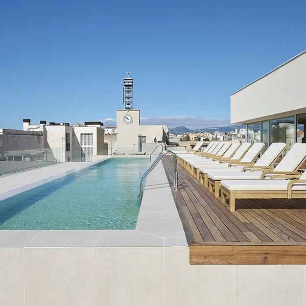 Dachschwimmbad mit klarem, blauem Wasser in MARINA CAMBRILS BY URH, gesäumt von weißen Liegestühlen auf einem Holzdeck, umgeben von modernen Gebäuden unter einem strahlend blauen Himmel.