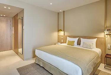 Marina Cambrils room Ein modernes Hotelzimmer im MARINA CAMBRILS BY URH verfügt über ein großes, sauber gemachtes Bett, neutrale Beige- und Cremetöne, zwei Kissen, ein kleines gestreiftes Kissen, Nachttischlampen und einen Spiegel neben der hölzernen Eingangstür.