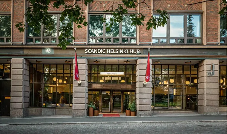 Der Eingang des Scandic Helsinki Hub, eines modernen Hotels mit großen Fenstern, roten Fahnen auf beiden Seiten der Tür und einem Schild über dem Eingang - ideal für Reisende, die von einer Golfrundreise nach Irland zurückkehren. Davor sind Bäume und eine Straße mit Kopfsteinpflaster zu sehen.