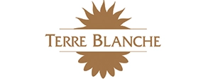 Logo für Terre Blanche in braunen Großbuchstaben, zentriert über einem weißen horizontalen Band mit einer Sonnenblume oder einem blumenähnlichen Muster im Hintergrund, perfekt für Golf- und Reiseliebhaber, die Eleganz suchen.