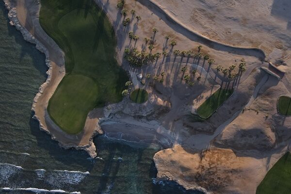 Luftaufnahme eines Golfplatzes am Meer mit grünen Fairways, Sandflächen, Palmen, die lange Schatten werfen, und Wellen, die an die Küste branden – ein Traumziel, das ganz oben auf Ihrer Packliste für eine Golfreise nach Ägypten stehen wird.