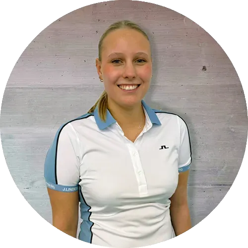 Eine Frau mit blondem Haar in einem Pferdeschwanz und einem weiß-hellblauen Sporthemd mit Kragen steht lächelnd vor einer strukturierten, hellen Wand.