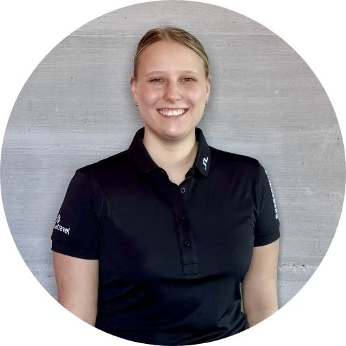 Darleen Wehner, mit hellen, zurückgebundenen Haaren, steht lächelnd in einem schwarzen Kragenhemd vor einer schlichten hellgrauen Betonwand.