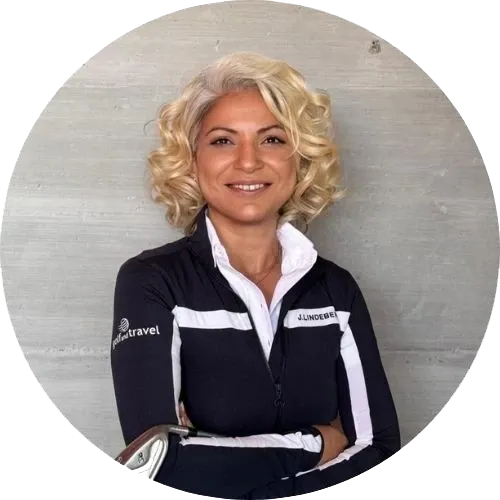 Eine Frau mit kurzen, lockigen blonden Haaren steht lächelnd und mit verschränkten Armen da. Sie trägt eine schwarz-weiße Sportjacke mit Logos an den Ärmeln vor einem schlichten grauen Hintergrund.