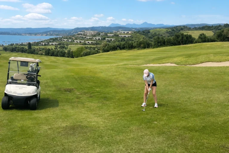 Eine Person macht sich bereit, auf einem grünen Fairway beim Golfen in Costa Navarino einen Golfball zu schlagen; daneben steht ein Golfwagen. Sanfte Hügel, Gebäude und ein blauer Himmel mit vereinzelten Wolken runden die malerische Kulisse ab.