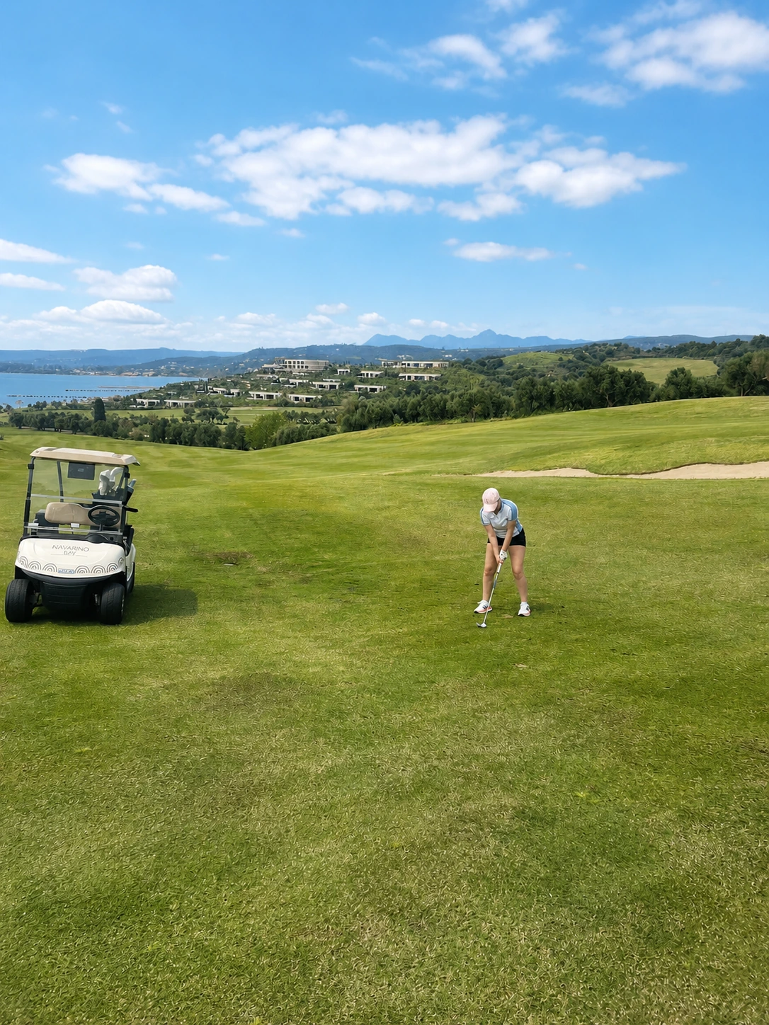 Navarino_Golf_800_or_original Eine Person macht sich bereit, auf einem grünen Fairway beim Golfen in Costa Navarino einen Golfball zu schlagen; daneben steht ein Golfwagen. Sanfte Hügel, Gebäude und ein blauer Himmel mit vereinzelten Wolken runden die malerische Kulisse ab.