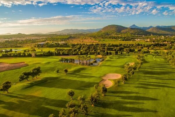 Luftaufnahme eines üppig bewachsenen Golfplatzes mit Sandbunkern, Wasserhindernissen, Bäumen und umliegenden Hügeln unter blauem Himmel mit vereinzelten Wolken – perfekt für eine Anfänger-Golfreise auf Mallorca.