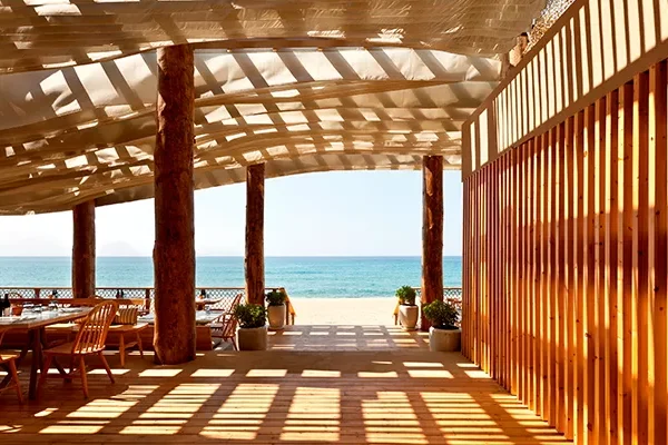 Eine sonnendurchflutete Terrasse unter freiem Himmel direkt am Strand mit Holzmöbeln und Säulen bietet einen Blick auf das Meer. Die Schatten des Lamellendachs zeichnen Muster auf den Boden, während man den Blick auf das blaue Meer genießen kann – perfekt nach einem Tag auf dem Golfplatz in Costa Navarino.