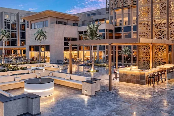 Moderner Loungebereich im Freien in einem Resort mit geschwungenen Sitzgelegenheiten, einer zentralen Feuerstelle, einer Bar mit Hockern, Palmen und dekorativen Holzparavents – ideal zum Entspannen nach einem Tag auf einer Golfreise nach Ägypten – umgeben von modernen Gebäuden in der Abenddämmerung.