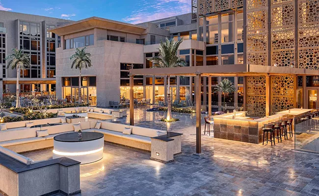 erfahrungsbericht-aegypten Moderner Loungebereich im Freien in einem Resort mit geschwungenen Sitzgelegenheiten, einer zentralen Feuerstelle, einer Bar mit Hockern, Palmen und dekorativen Holzparavents – ideal zum Entspannen nach einem Tag auf einer Golfreise nach Ägypten – umgeben von modernen Gebäuden in der Abenddämmerung.