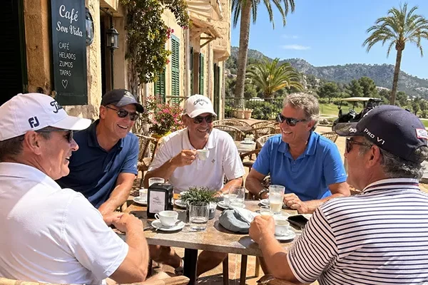 Fünf Männer sitzen um einen Tisch in einem Café im Freien, lächeln und unterhalten sich. Sie tragen legere Golfkleidung und Sonnenbrillen. Im Hintergrund sind Palmen und ein Golfplatz an einem sonnigen Tag zu sehen.