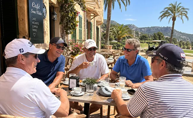 erfahrungsbericht-golfen-mallorca Fünf Männer sitzen um einen Tisch in einem Café im Freien, lächeln und unterhalten sich. Sie tragen legere Golfkleidung und Sonnenbrillen. Im Hintergrund sind Palmen und ein Golfplatz an einem sonnigen Tag zu sehen.