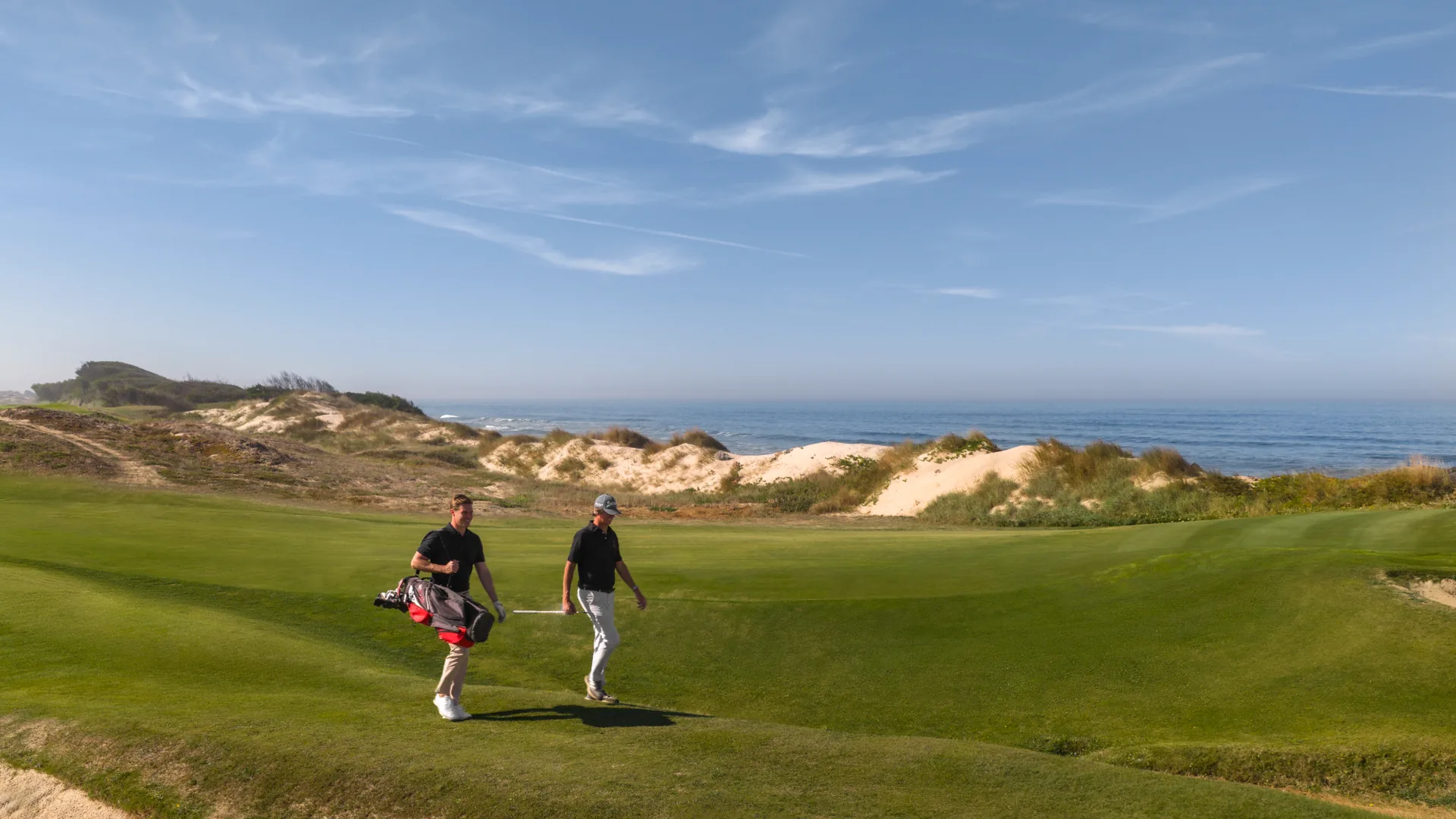 Zwei Personen spazieren entlang eines üppig grünen Golfplatzes in der Nähe von Sanddünen, eine trägt eine Golftasche - eine idyllische Szene für alle, die Golf und Reisen lieben. Der Ozean und der blaue Himmel mit Wolkenfetzen bieten eine atemberaubende Kulisse.
