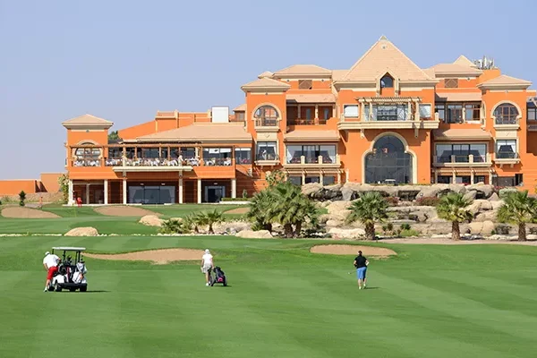 Golfer spielen auf einem grünen Fairway vor einem großen, orangefarbenen Clubhaus im mediterranen Stil mit Balkonen und Palmen unter strahlend blauem Himmel – eine Szene, die an einen Erfahrungsbericht einer Golfreise nach Ägypten erinnert.