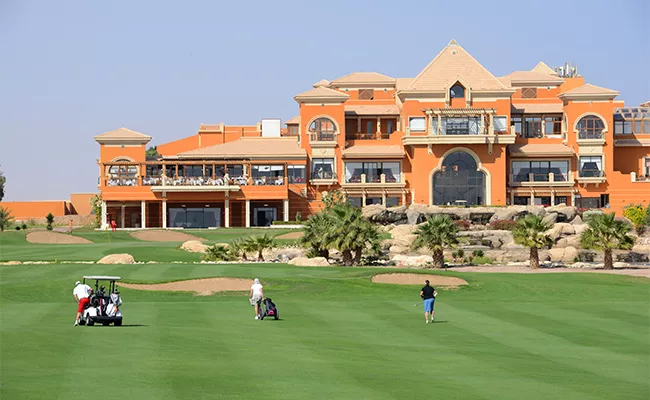 golf-anfaengerreise-aegypten Golfer spielen auf einem grünen Fairway vor einem großen, orangefarbenen Clubhaus im mediterranen Stil mit Balkonen und Palmen unter strahlend blauem Himmel – eine Szene, die an einen Erfahrungsbericht einer Golfreise nach Ägypten erinnert.