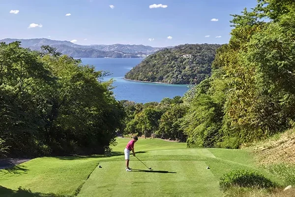 Ein Golfer bereitet sich darauf vor, auf einem üppig grünen, von Bäumen umgebenen Platz abzuschlagen, mit einem malerischen Blick auf Hügel und einen blauen See oder das Meer im Hintergrund unter einem klaren Himmel.