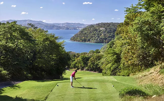 Ein Golfer bereitet sich darauf vor, auf einem üppig grünen, von Bäumen umgebenen Platz abzuschlagen, mit einem malerischen Blick auf Hügel und einen blauen See oder das Meer im Hintergrund unter einem klaren Himmel.