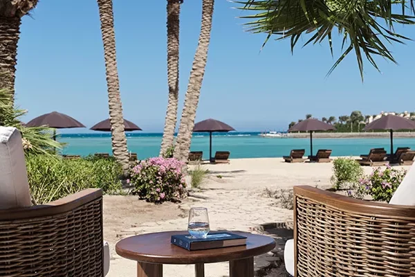 Zwei Korbstühle und ein kleiner Tisch mit einem Glas Wasser und einem Buch bieten einen Blick auf einen Sandstrand, das türkisfarbene Meer, Palmen und Sonnenliegen – ideal, um an einem sonnigen Tag einen Erfahrungsbericht über eine Golfreise nach Ägypten zu lesen.