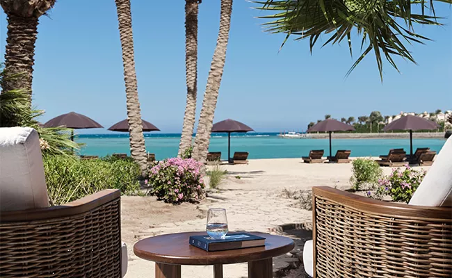 golf-gruppenreise-aegypten-2 Zwei Korbstühle und ein kleiner Tisch mit einem Glas Wasser und einem Buch bieten einen Blick auf einen Sandstrand, das türkisfarbene Meer, Palmen und Sonnenliegen – ideal, um an einem sonnigen Tag einen Erfahrungsbericht über eine Golfreise nach Ägypten zu lesen.