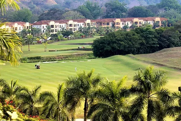 Ein üppig bewachsener Golfplatz, auf dem Menschen spielen, umgeben von Palmen und viel Grün, mit einer Reihe von Gebäuden im mediterranen Stil und Hügeln im Hintergrund.