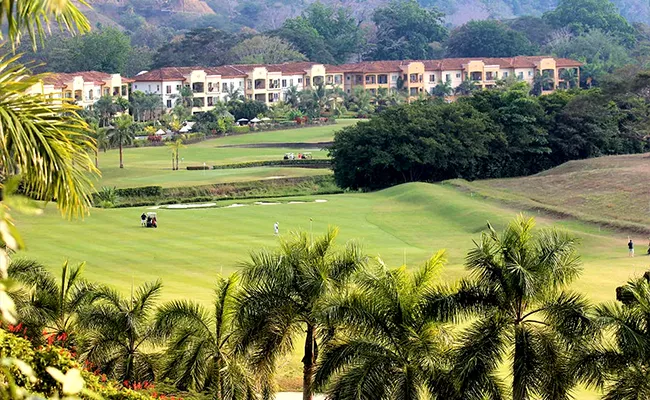 Ein üppig bewachsener Golfplatz, auf dem Menschen spielen, umgeben von Palmen und viel Grün, mit einer Reihe von Gebäuden im mediterranen Stil und Hügeln im Hintergrund.