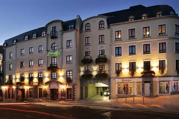 Ein vierstöckiges, cremefarbenes Hotelgebäude mit Bogenfenstern und Balkonen ist in der Abenddämmerung in warmes Licht getaucht; die Geschäfte im Erdgeschoss und ein Torbogen am Eingang sind zu erkennen und erinnern an Boutique-Hotels in der Nähe berühmter Golfplätze in Irland. Lichtspuren vorbeifahrender Autos säumen die Straße davor.