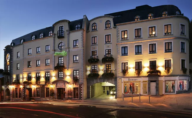 golf-gruppenreise-irland-2 Ein vierstöckiges, cremefarbenes Hotelgebäude mit Bogenfenstern und Balkonen ist in der Abenddämmerung in warmes Licht getaucht; die Geschäfte im Erdgeschoss und ein Torbogen am Eingang sind zu erkennen und erinnern an Boutique-Hotels in der Nähe berühmter Golfplätze in Irland. Lichtspuren vorbeifahrender Autos säumen die Straße davor.