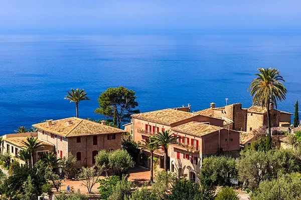 Steinhäuser im mediterranen Stil mit roten Fensterläden und Ziegeldächern liegen inmitten von Palmen und bieten einen Blick auf das ruhige blaue Meer – die perfekte Kulisse für eine Golf-Gruppenreise auf Mallorca unter strahlend blauem Himmel.