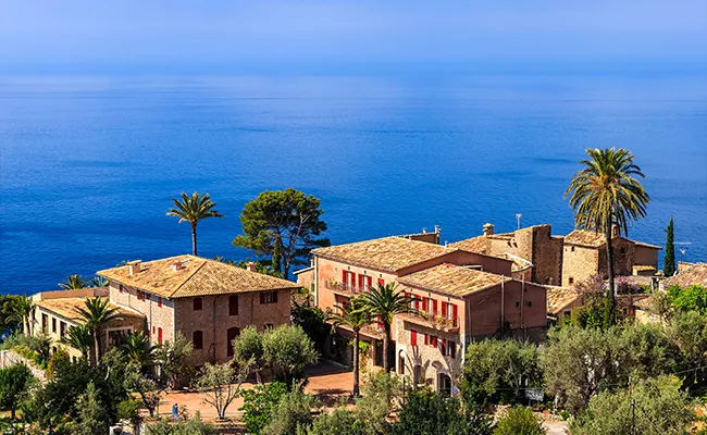 golf-gruppenreise-mallorca-2 Steinhäuser im mediterranen Stil mit roten Fensterläden und Ziegeldächern liegen inmitten von Palmen und bieten einen Blick auf das ruhige blaue Meer – die perfekte Kulisse für eine Golf-Gruppenreise auf Mallorca unter strahlend blauem Himmel.