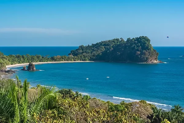 golf-reisebericht-costa-rica Ein tropischer Strand mit türkisfarbenem Wasser, üppig grünen Bäumen und einer kleinen, bewaldeten Insel vor der Küste unter einem strahlend blauen Himmel. Auf dem Wasser nahe der Küste sind mehrere kleine Boote zu sehen.