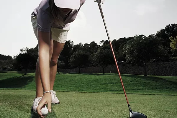 Ein Golfer mit Mütze und Shorts beugt sich vor, um einen Golfball auf das Tee zu legen, während er einen Golfschläger in der Hand hält – im Hintergrund sind üppige Bäume zu sehen – perfekt für eine entspannte Golf-Gruppenreise nach Mallorca.