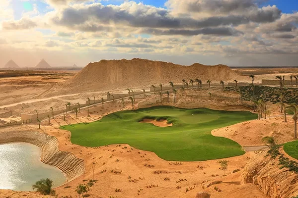 Ein üppig begrünter Golfplatz mit einem Wasserhindernis liegt inmitten einer Wüstenlandschaft. Sanddünen und Palmen umgeben den Platz, und in der Ferne sind die Pyramiden von Gizeh zu sehen – perfekt für einen unvergesslichen Erfahrungsbericht über eine Golfreise nach Ägypten.