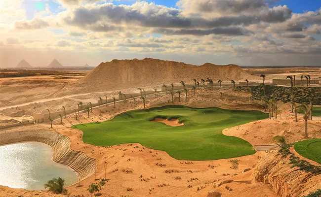 golfplaetze-aegypten Ein üppig begrünter Golfplatz mit einem Wasserhindernis liegt inmitten einer Wüstenlandschaft. Sanddünen und Palmen umgeben den Platz, und in der Ferne sind die Pyramiden von Gizeh zu sehen – perfekt für einen unvergesslichen Erfahrungsbericht über eine Golfreise nach Ägypten.