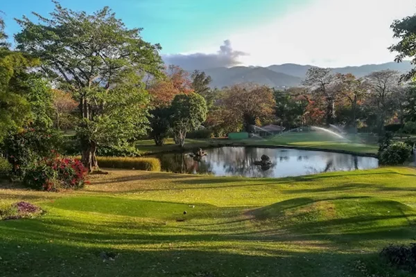 golfplaetze-costa-rica Ein üppig grüner Park mit einem kleinen Teich, umgeben von Bäumen und blühenden Sträuchern, mit Bergen im Hintergrund und einem Sprinkler, der den Rasen unter einem strahlenden, teilweise bewölkten Himmel bewässert.