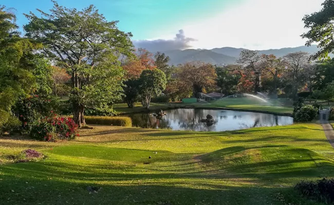 golfplaetze-costa-rica Ein üppig grüner Park mit einem kleinen Teich, umgeben von Bäumen und blühenden Sträuchern, mit Bergen im Hintergrund und einem Sprinkler, der den Rasen unter einem strahlenden, teilweise bewölkten Himmel bewässert.