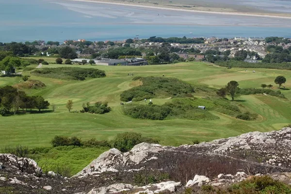 Ein malerischer Blick auf Golfplätze in Irland mit üppigen Grünflächen, Sanddünen, Bäumen und einigen in der Ferne liegenden Gebäuden in der Nähe einer sandigen Küste und des blauen Meeres, alles gesehen von einem felsigen Hügel aus.