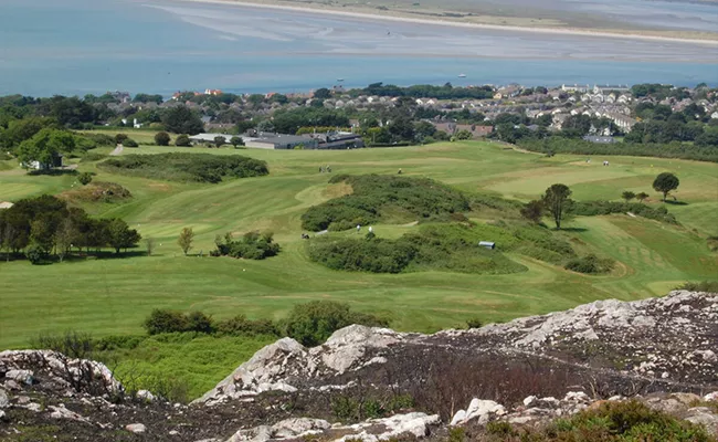 Ein malerischer Blick auf Golfplätze in Irland mit üppigen Grünflächen, Sanddünen, Bäumen und einigen in der Ferne liegenden Gebäuden in der Nähe einer sandigen Küste und des blauen Meeres, alles gesehen von einem felsigen Hügel aus.